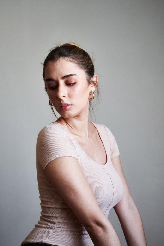 Marintia Bustos a model with a radiant complexion poses gracefully against a neutral backdrop. She wears a soft pink top with a scoop neckline and gold hoop earrings. Her gaze is downward and her posture exudes confidence.